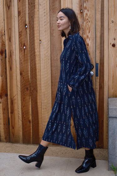 A woman standing in front of a wooden gate wearing black ikat shirt dress with long sleeves.