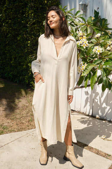 Woman wearing a long white dress with a slit, standing outdoors near a white building and greenery.