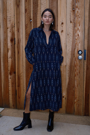 A woman standing in front of a wooden gate wearing black ikat shirt dress with long sleeves.
