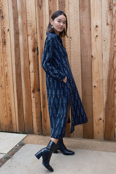 A woman standing in front of a wooden gate wearing black ikat shirt dress with long sleeves.