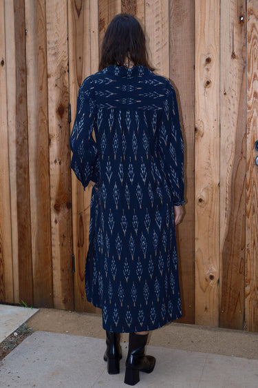 A woman standing in front of a wooden gate wearing black ikat shirt dress with long sleeves, back view.