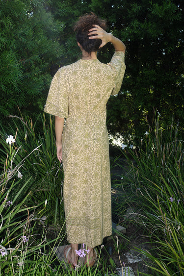 woman standing in garden wearing a light green print tapestry dress with zip front, v neck and elbow length sleeves, back view.