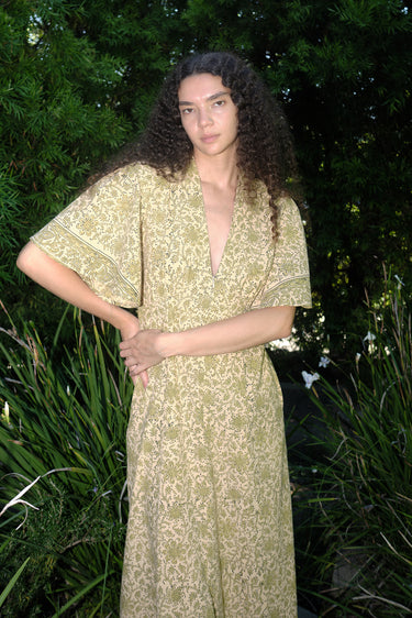 woman standing in garden wearing a light green print tapestry dress with zip front, v neck and elbow length sleeves