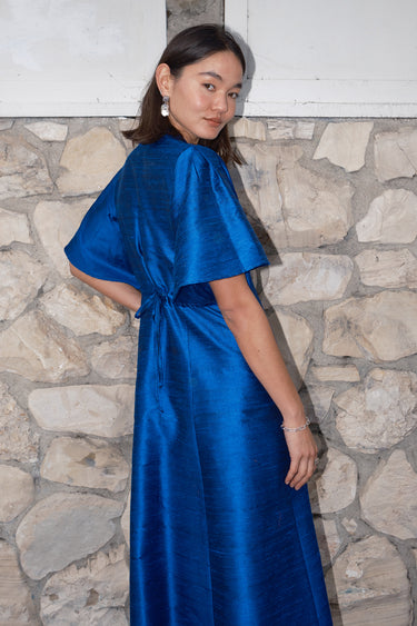 woman standing by stone wall wearing a sapphire blue dress . Back view