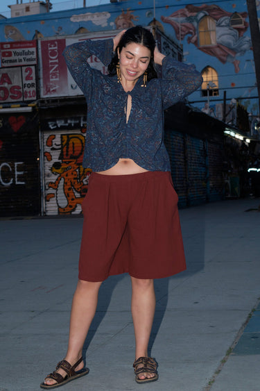 A woman standing on the sidewalk wearing a raisin colored Bermuda short, long sleeve top and a pair of strappy sandals. The background includes graffiti and a building.