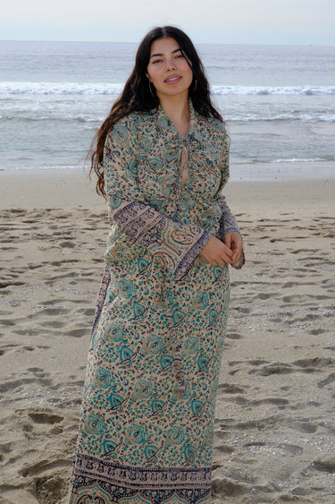 A woman standing at the beach wearing tan with green turquoise flower tapestry shirt with bell sleeves and a matching skirt.