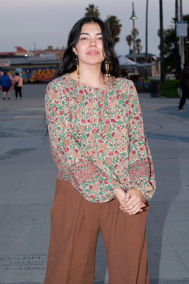 A woman standing at a beach boardwalk wearing a floral print tapestry blouse and a nutmeg brown trouser.