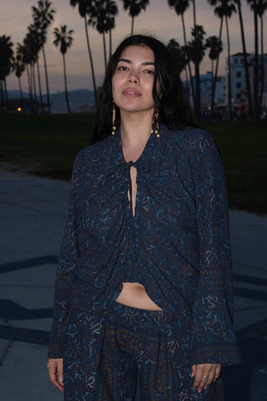 A woman standing at Venice Beach at dusk wearing dark blue midnight sky handloom tapestry shirt with bell sleeves and matching trousers.