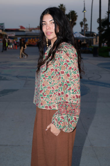 A woman standing at a beach boardwalk wearing a nutmeg brown trouser and a floral print blouse. Side View