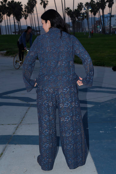 A woman standing at Venice Beach at dusk wearing dark blue midnight sky handloom tapestry low rise trouser and a matching shirt with bell sleeves. Back View