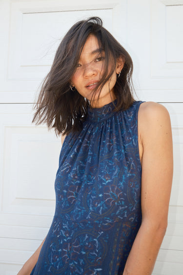 A woman standing in front of a white garage door, wearing a navy dress with a band collar, empire waist, and hand woven, hand block printed Indian Tapestry fabric.