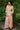 Woman wearing a gold top and pink skirt standing in front of green foliage.