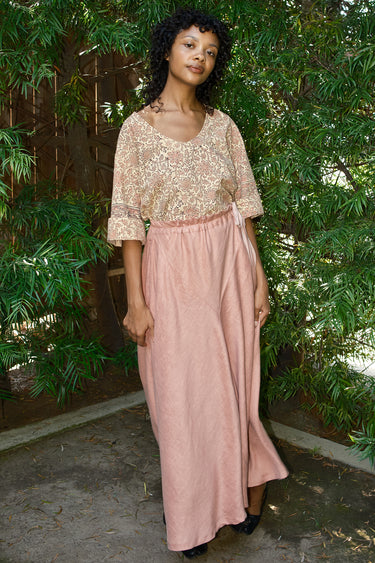 Woman wearing a tapestry top and pink skirt standing outdoors with greenery in the background