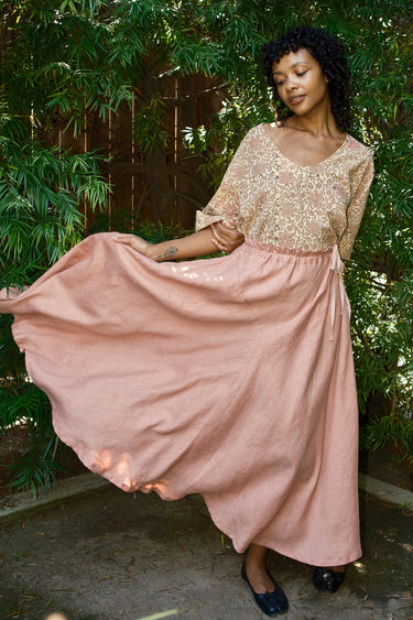 Woman in a gold top and pink skirt standing outdoors with greenery in the background