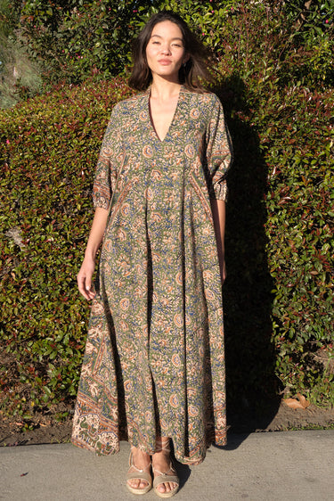 A woman wearing a maxi caftan with a floral pattern in a color that could be described as celadon green ground with tan, brown and blue, standing outdoors.