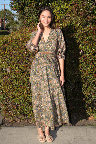 A woman wearing a maxi caftan with a floral pattern in a color that could be described as celadon green ground with tan, brown and blue, belted, standing outdoors.