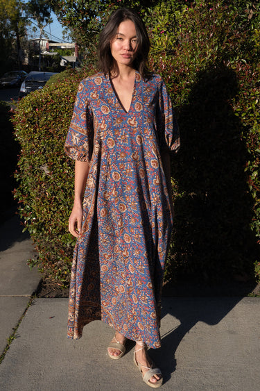 A woman wearing a maxi caftan with a floral pattern in a color that could be described as blue ground with tan, brown and coral, standing outdoors.