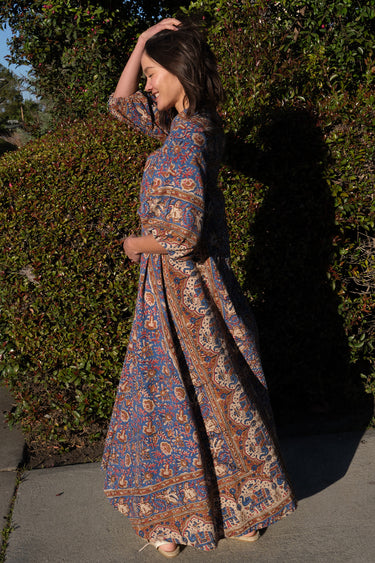 A person wearing a traditional blue caftan with a paisley pattern and a belt, standing outdoors.