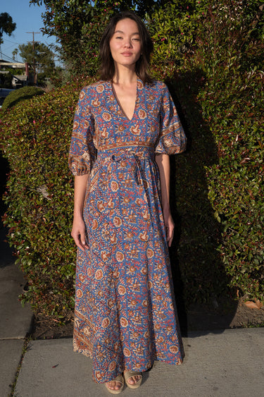 A woman wearing a maxi caftan with a floral pattern in a color that could be described as blue ground with tan, brown and coral, belted, standing outdoors.