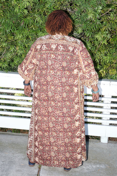 A woman outside standing against greenery wearing a raisin floral tapestry maxi caftan with three quarter balloon sleeves, back view.