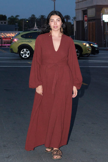 A woman standing outside in the street wearing a raisin osnaburg maxi caftan with long full sleeves and a belt.