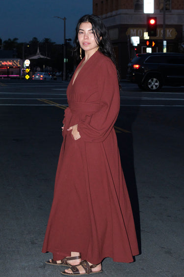 A woman standing outside in the street wearing a raisin osnaburg maxi caftan with long full sleeves and a belt ,side view.