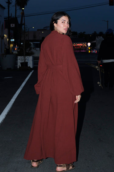 A woman standing outside in the street wearing a raisin osnaburg maxi caftan with long full sleeves and a belt, back view.