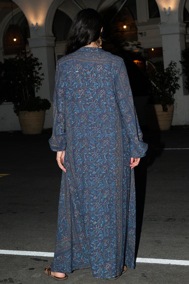A woman standing outside a night wearing a long sleeve maxi caftan and sandals. The caftan is cut in midnight sky handloom tapestry, back view.
