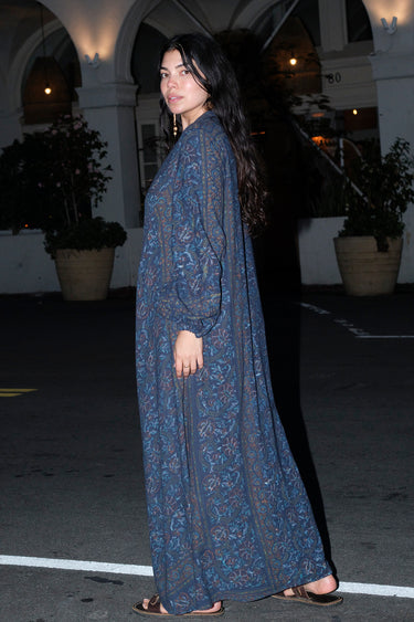 A woman standing outside a night wearing a long sleeve maxi caftan and sandals. The caftan is cut in midnight sky handloom tapestry, side view.
