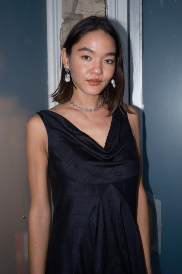 A woman standing on a doorway wearing a sleeveless, full-length black dress with a full drape neckline, side zipper, and pockets.