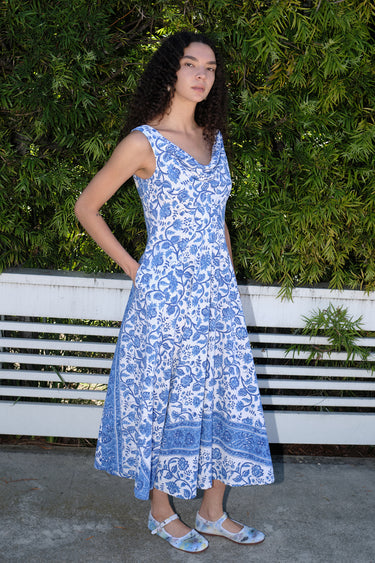 Woman standing in front of white fence and hedge wearing a white with blue tapestry midi dress with a draped neck and very full skirt.
