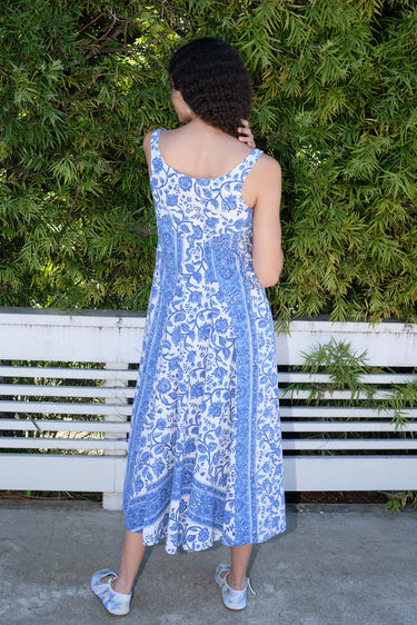 Woman standing in front of white fence and hedge wearing a white with blue tapestry midi dress with a draped neck and very full skirt, back view.
