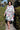 A woman standing on a sidewalk wearing a white coat dress with a colorful floral tapestry pattern, featuring patch pockets and a tie belt.
