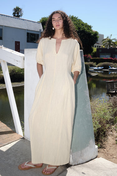 A woman standing outside by a canal wearing an alabaster off white maxi dress with shape from princess panels.