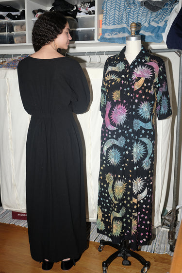 A woman standing inside wearing a black maxi dress with shape from princess panels, back view.
