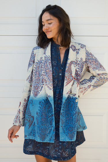 A woman smiling, wearing a double-breasted jacket with an indigo and blue pattern jacket, over a dress in a dark blue tapestry pattern. The jacket is dipped in indigo dye and has pockets on the inside.