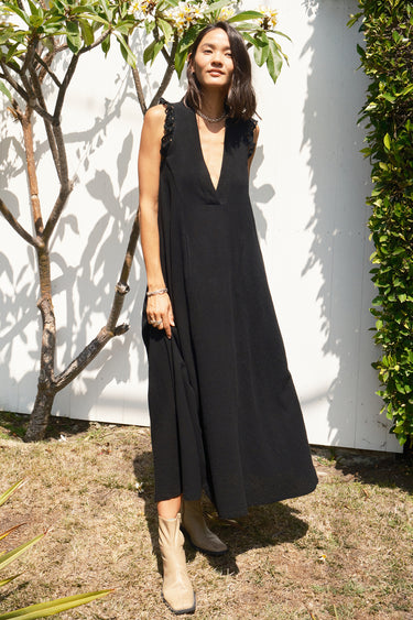 Woman wearing a black sleeveless dress - caftan standing outdoors against a white wall.