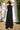 Woman wearing a black sleeveless dress - caftan standing outdoors against a white wall. Back view