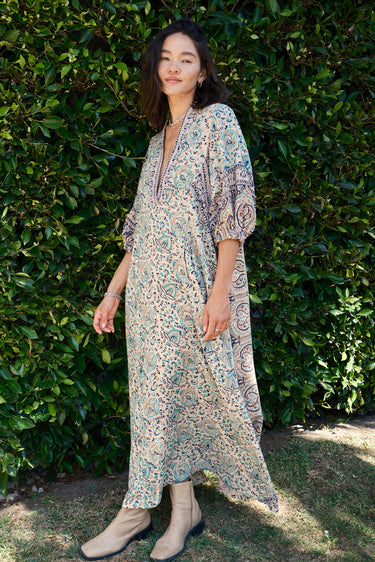 A female model wearing a multicolored floral caftan with three-quarter sleeves and standing in front of a hedge.