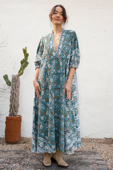 Woman wearing a long, patterned caftan standing against a white wall with a cactus plant.