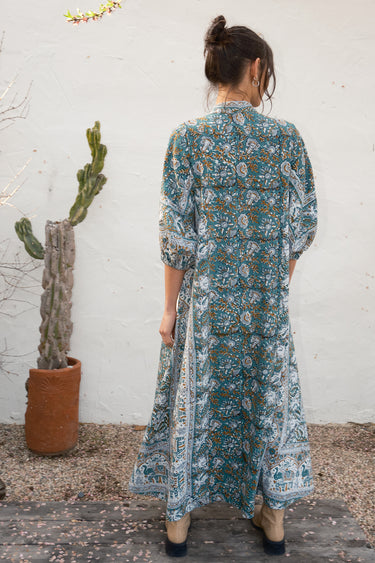 Person wearing a patterned caftan in a rustic setting with a cactus in the background.