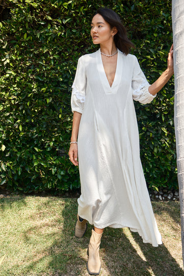 A woman standing outdoors wearing a white caftan with 3/4 balloon sleeves and a deep 'V' neckline. The caftan has pockets on the front panel and is made of a non-sheer crinkle textured fabric.