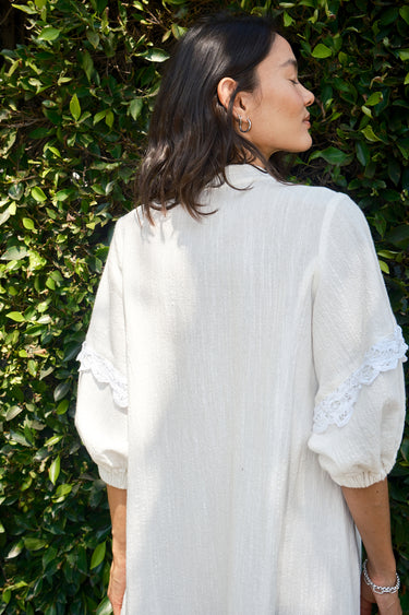 A woman standing outdoors wearing a white caftan with 3/4 balloon sleeves and a deep 'V' neckline. The caftan has pockets on the front panel and is made of a non-sheer crinkle textured fabric. Back view