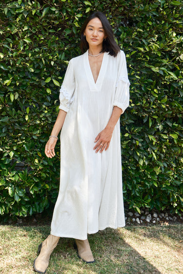 A woman standing outdoors wearing a white caftan with 3/4 balloon sleeves and a deep 'V' neckline. The caftan has pockets on the front panel and is made of a non-sheer crinkle textured fabric.