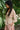 Woman wearing a patterned top and pink skirt standing among green foliage.
