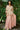 Woman wearing a beige top and pink skirt standing in front of green foliage.