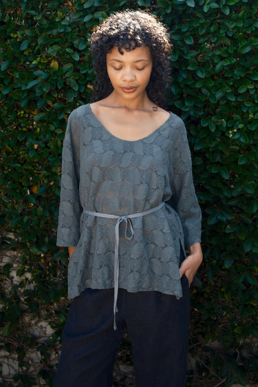 Woman wearing a gray patterned top and dark pants against a green leafy background