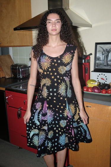 A woman standing in a kitchen wearing a knee-length dress in a black ground tapestry with rainbow suns, moons and stars printed on it.