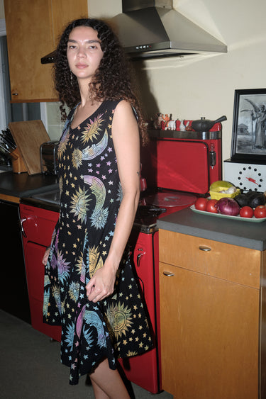 A woman standing in a kitchen wearing a knee-length dress in a black ground tapestry with rainbow suns, moons and stars printed on it.