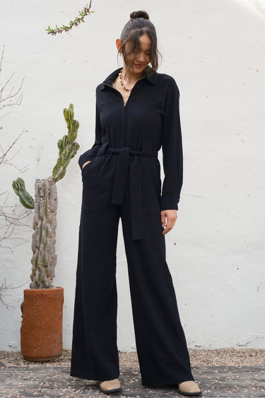 Woman wearing a black jumpsuit standing against a white wall with a plant in the background